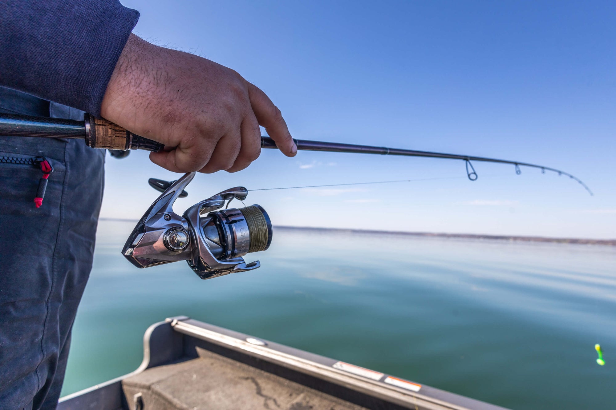 A hand is holding onto a fishing pole that is lined with Cortland Master Braid Bronze. There is a lure at the end of the line and water in the background.