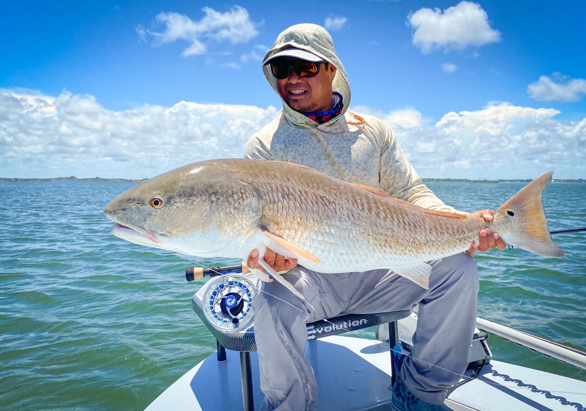A fisherman is seated on a boat, while holding up a large redfish.