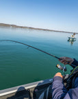 A man is overlooking the water, as he is reeling in his pole. There is another person standing on a boat in the background. 
