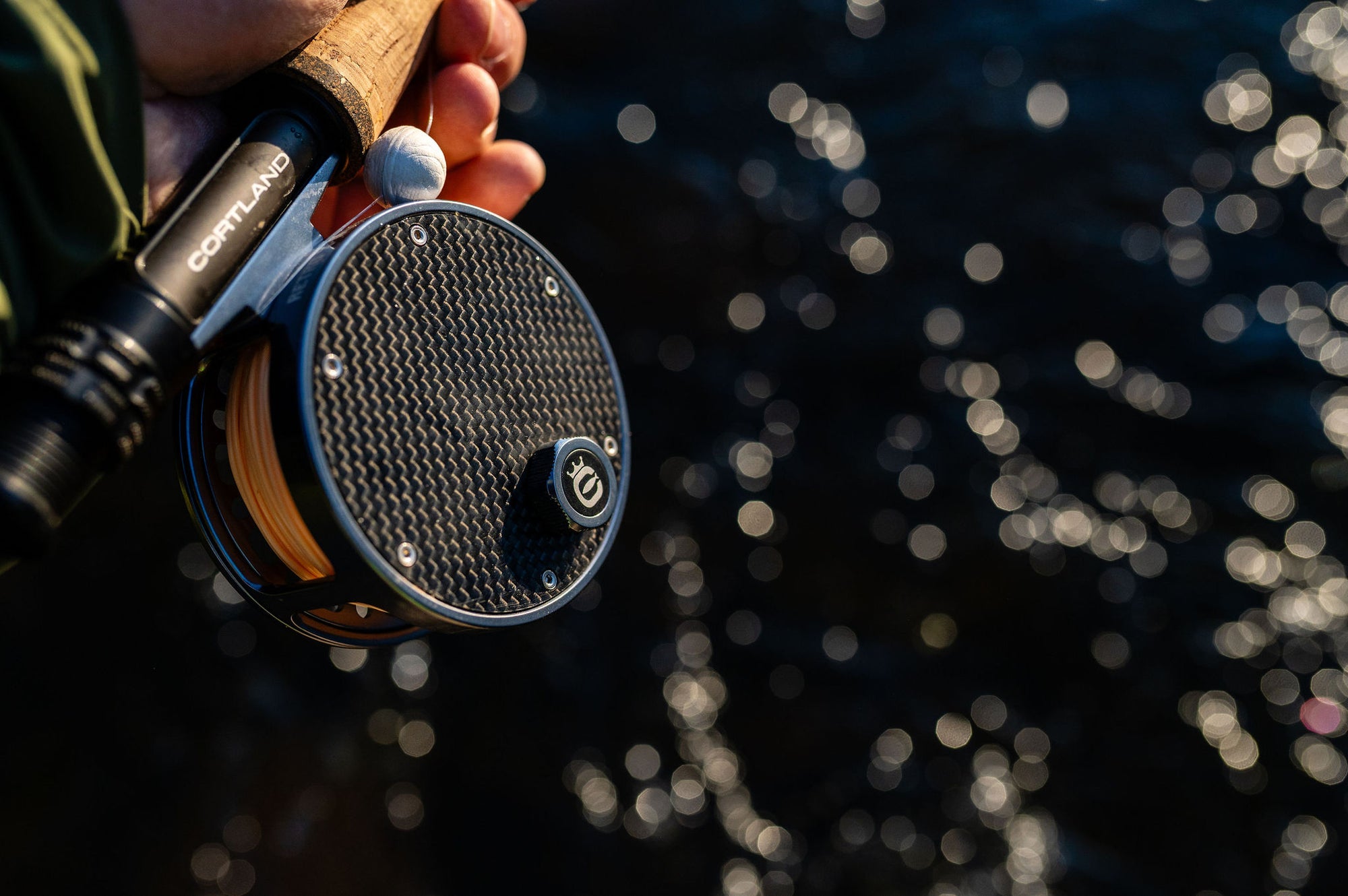 Close-up of a fly fishing reel with a carbon fiber patterned spool, held by a person whose hand is visible, with a blurred background of sparkling water.