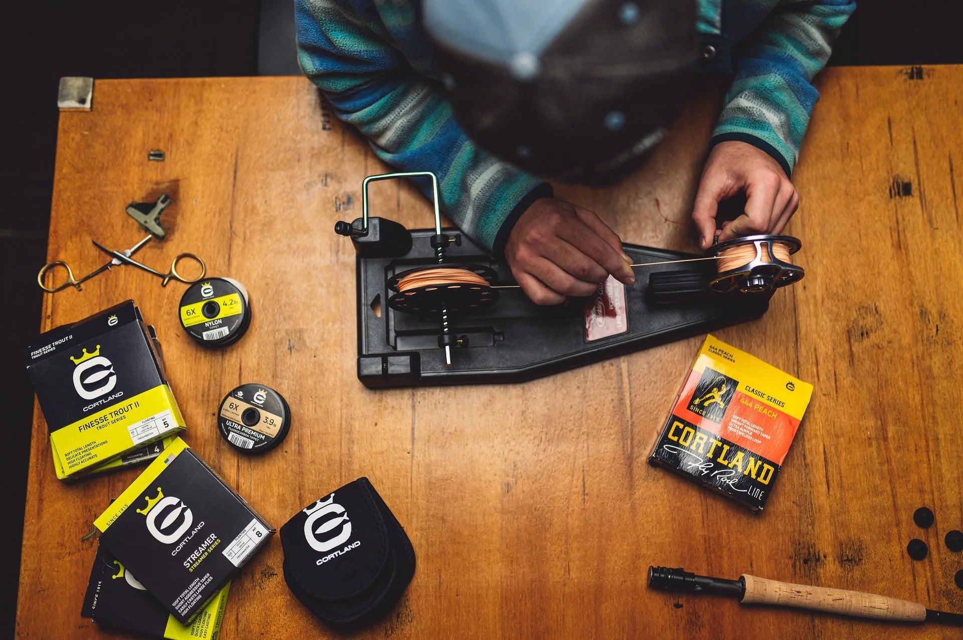 Person working on a fishing reel with various fishing equipment on a wooden table
