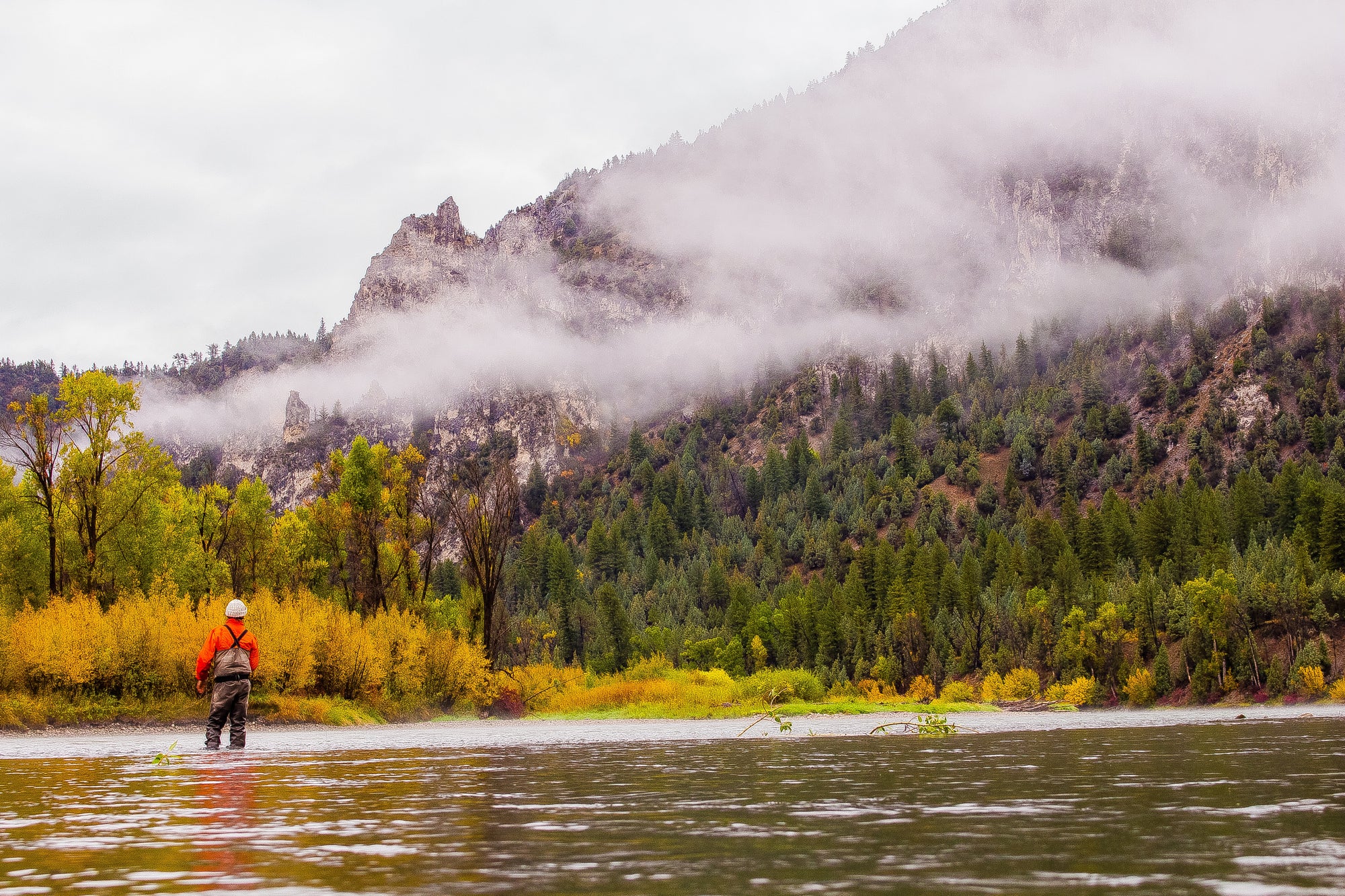 Someone standing in knee high water with their back to the camera. In the background there is a mountain and fog is rolling in with trees on the lefthand side. 