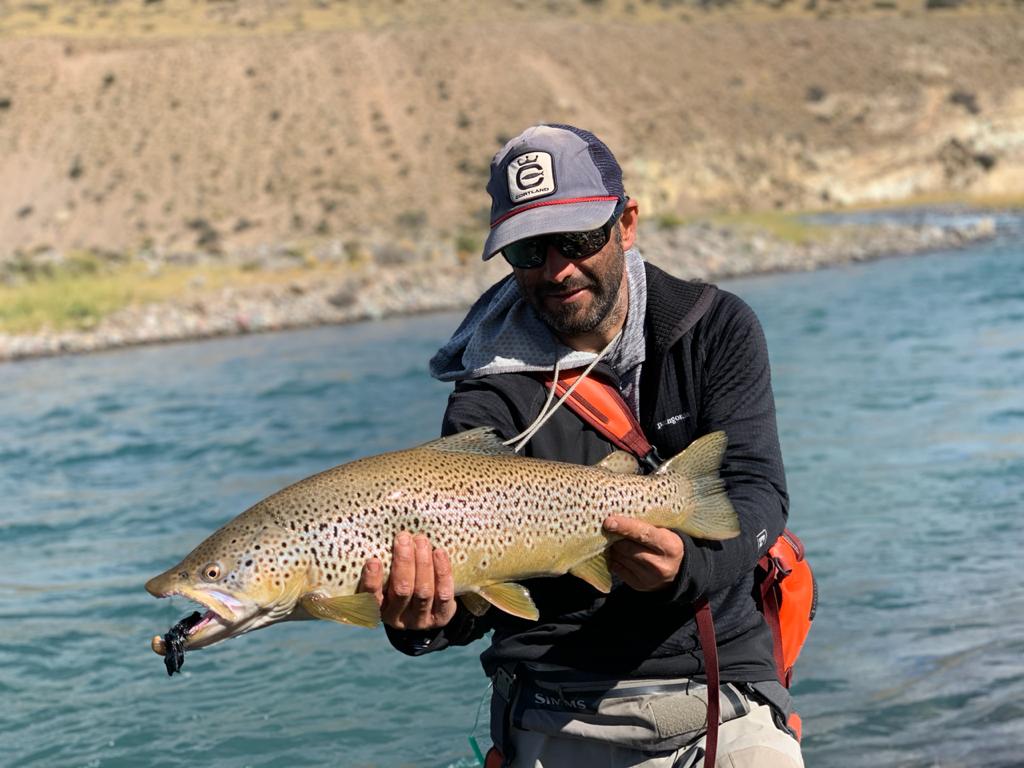 A fisherman is holding a trout with a black lure in its mouth. You can see water and a hill in the background