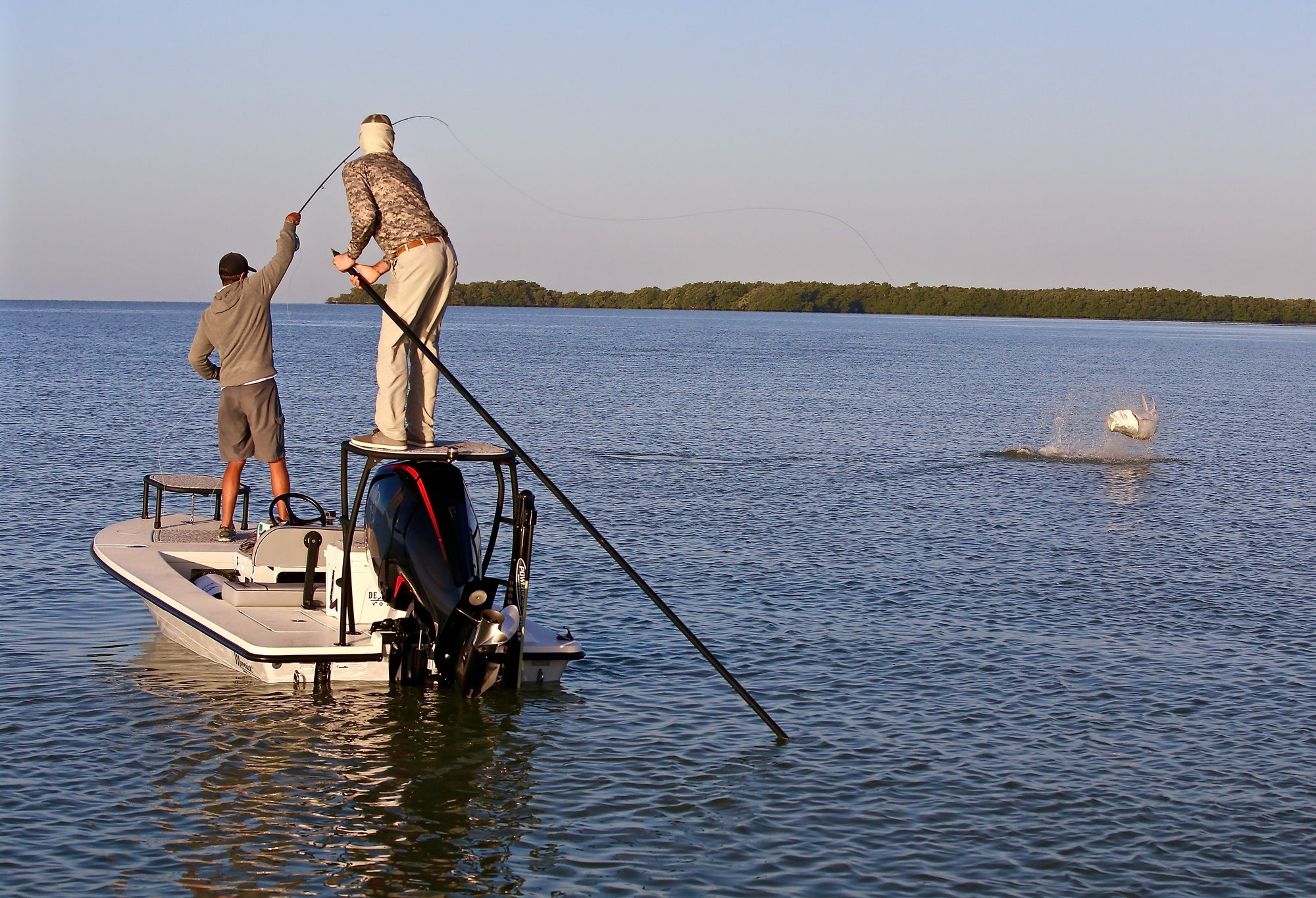 Two anglers fishing on a boat. One is holding onto his fishing pole, while a fish is jumping out of the water. 